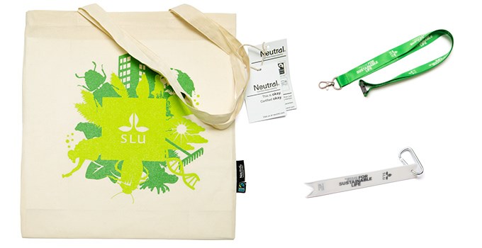 The SLU tote bag in off white with a green SLU brand print, a green key strap with a SLU logo and a white clip-on reflector with a black SLU logo and brand promise. Photo.