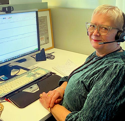 En person sitter vid ett skrivbord och arbetar på en dator. Personen bär ett headset och har korsade händer på skrivbordet.