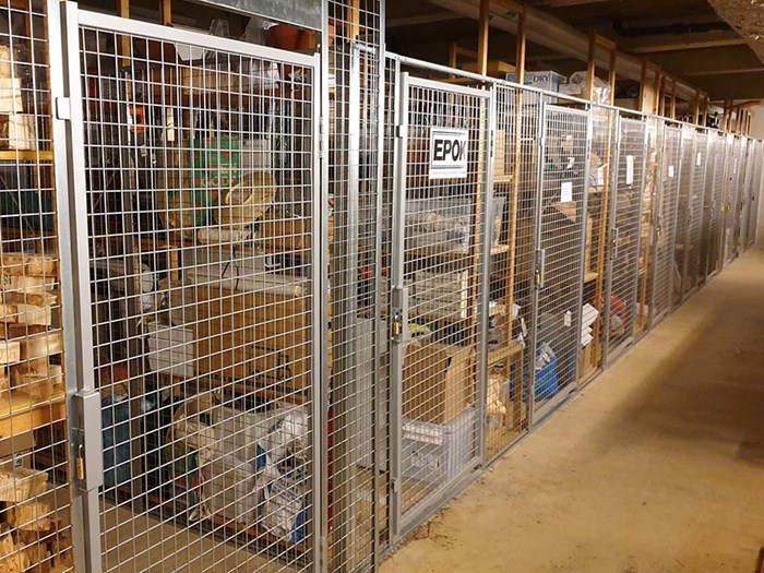 Bild av ett inomhus förrådsutrymme med metallstängsel som separerar olika förrådsenheter fyllda med lådor och diverse föremål, foto.