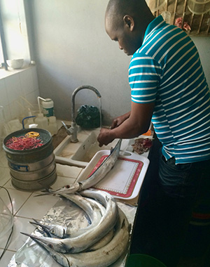 A man standing at a sink and rinsing off something under a tap. Beside him are fishes on two trays. Photo.
