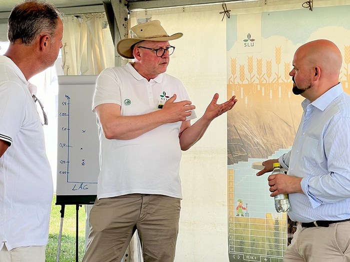 Tre personer står i ett tält och verkar vara involverade i en diskussion eller presentation om jordbruk, med en whiteboard och en affisch som visar vete och grafisk information i bakgrunden.