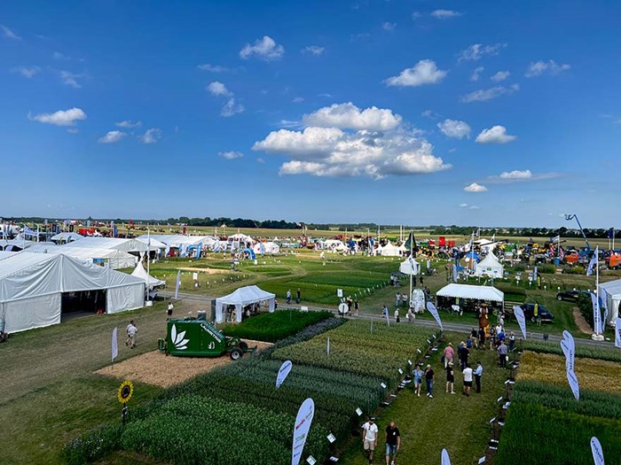 En bild av ett utomhusevenemang med flera vita tält och flaggor i ett stort öppet fält under en klarblå himmel. Det finns olika grödor som växer i rader och många människor spridda över platsen. Evenemanget verkar vara en lantbruksmässa eller utställning.