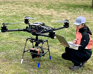 Sittande kvinna bredvid stor drönare på gräsmatta. Foto.