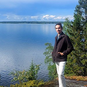 Félix Barbut wearing a white trousers and a dark jacket is standing in front of a lake in a typical Northern Swedish landscape