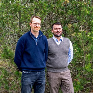 Two men are standing with the arms crossed behind the back in front of a green bush