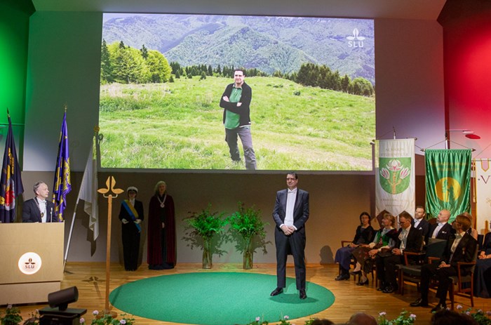 A festively dressed man stands in the middle of a decorated stage. Behind him a picture of a man standing in nature among green hills is projected on the wall.