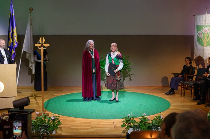 Two persons on a stage on a green carpet. One of them with a big chain around her neck and a red robe, the other one wearing a traditional Swedish folk costume.